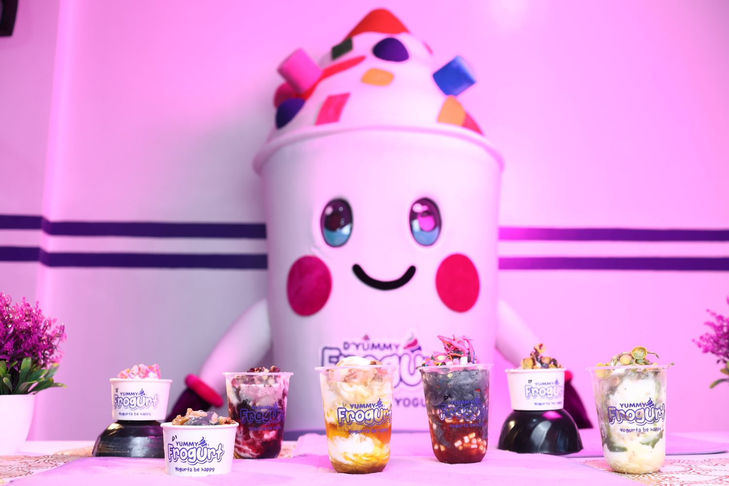 D'Yummy Frogurt mascot beside a spread of frozen yogurt cups